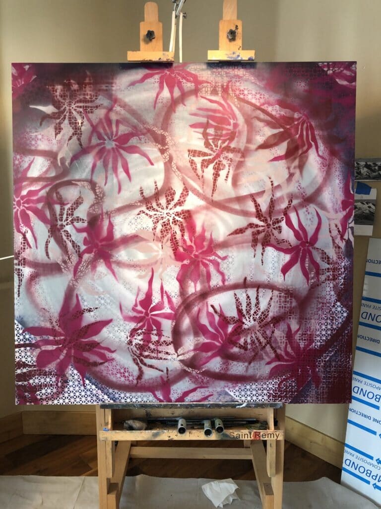 Acrylic spray painted base painting with spidery, deep pink and burgundy flowers float over a field of indigo/white grid pattern with abstract swirls through out the panel. Set on an easel.