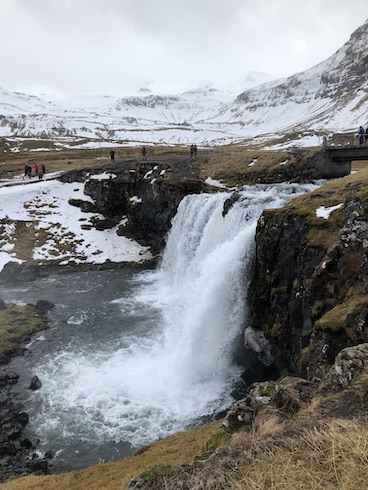 Photo of a beautiful waterfall with a drop of about 50 feet in the Iceland spring. Snow covered mountains surround this peaceful place, the warm, brown earth surrounding the falls.