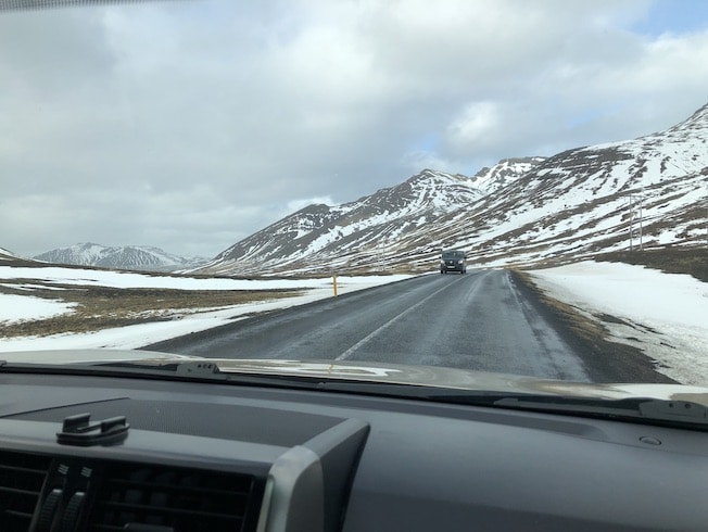 In this photo the viewer looks over the dashboard of the truck we are driving toward the snow spotted mountains that seem to slide up and out of the earth. An oncoming truck approaches on this cloudy, grey day.