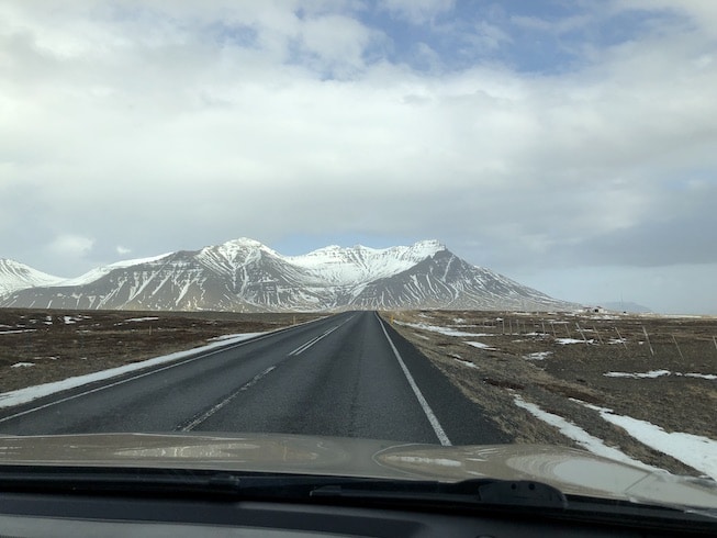 Photo of a long, straight road going straight as an arrow to the volcanic mountains of Iceland in the early spring. Snow still on the peaks but not much on the sides of the road