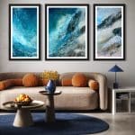 Ascent triptych by Cynthia McLoughlin showcased in a contemporary living space with light walls. The three abstract paintings, each in a black frame, depict swirling blue, turquoise and white forms reminiscent of mountain peaks. The artwork is mounted above a beige curved sofa with orange pillows complemented by a blue rug and modern decor.