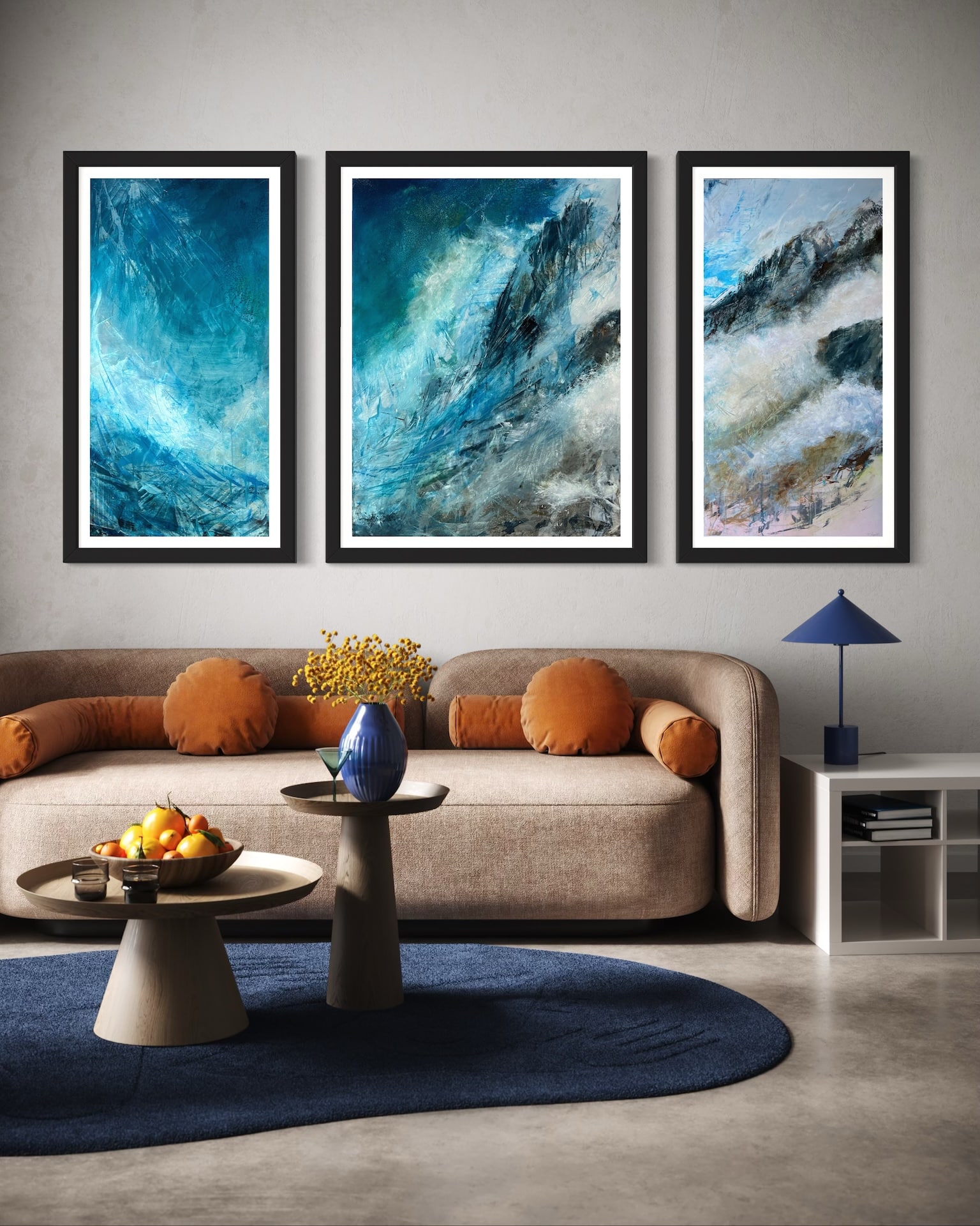 Ascent triptych by Cynthia McLoughlin showcased in a contemporary living space with light walls. The three abstract paintings, each in a black frame, depict swirling blue, turquoise and white forms reminiscent of mountain peaks. The artwork is mounted above a beige curved sofa with orange pillows complemented by a blue rug and modern decor.