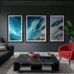 Ascent triptych by Cynthia McLoughlin displayed in a modern living room with dark walls. The three abstract panels feature dynamic blue, teal, and white brushstrokes, evoking the movement of mist over mountains. The artwork is framed in black and hans above a black marble coffee table, with a red accent chair and green plant nearby.